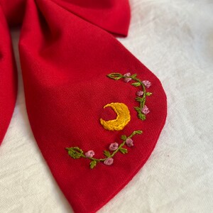 K&ouml;nnte beinhalten: Rote Stoff-Fliege mit einem gelben Halbmond und gr&uuml;nen Ranken mit rosa Bl&uuml;ten, die auf den Stoff gestickt sind.