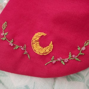 K&ouml;nnte beinhalten: Ein roter Stoff-Patch mit einer gelben gestickten Mondsichel und gr&uuml;nen gestickten Ranken mit rosa Bl&uuml;ten.