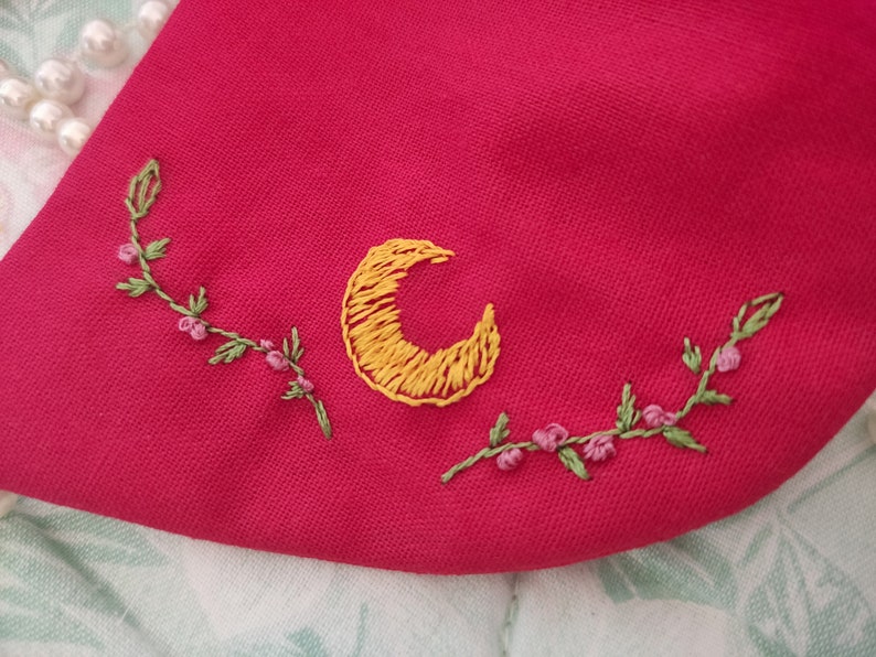K&ouml;nnte beinhalten: Ein roter Stoff-Patch mit einer gelben gestickten Mondsichel und gr&uuml;nen gestickten Ranken mit rosa Bl&uuml;ten.