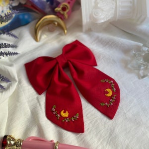 K&ouml;nnte beinhalten: Ein roter Fliege mit gestickten gelben Halbmonden und rosa Blumen. Die Fliege liegt auf einer wei&szlig;en Oberfl&auml;che.