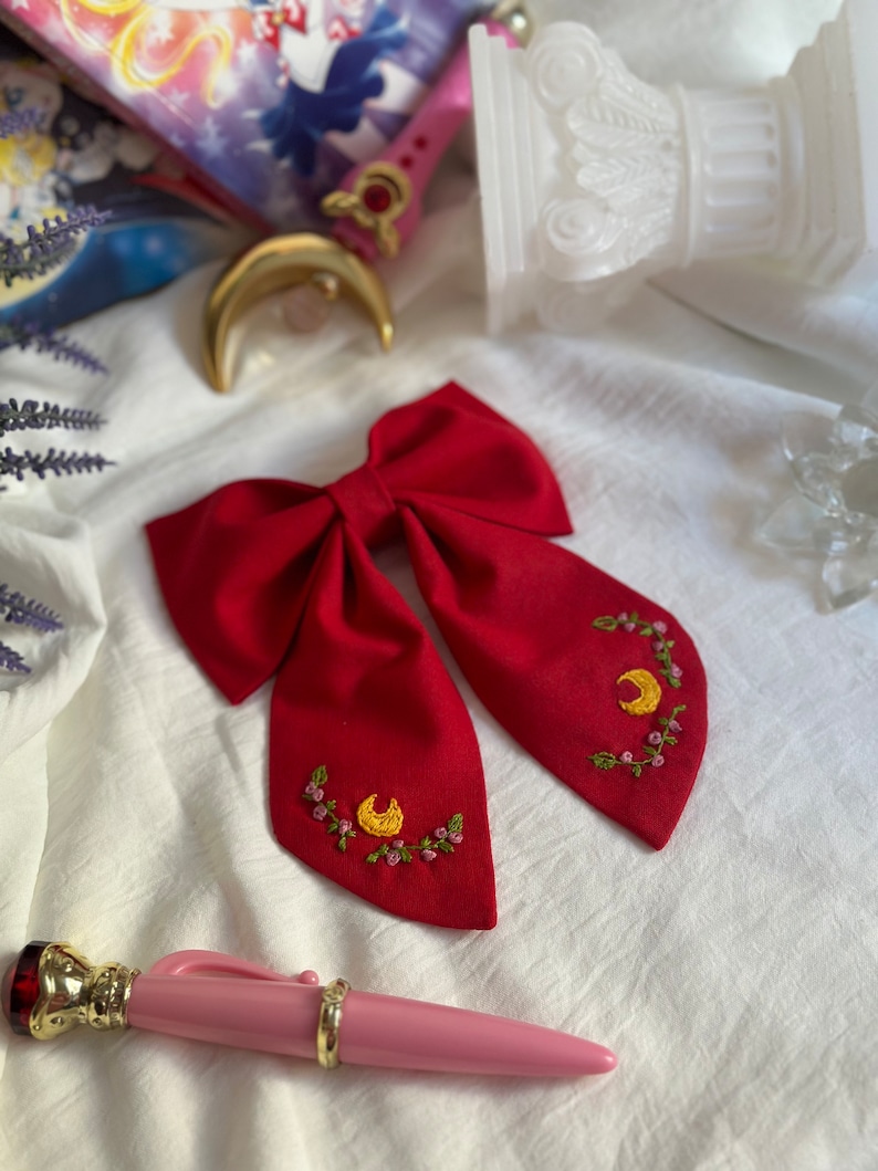 K&ouml;nnte beinhalten: Ein roter Fliege mit gestickten gelben Halbmonden und rosa Blumen. Die Fliege liegt auf einer wei&szlig;en Oberfl&auml;che.