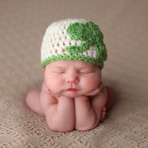 May include: A newborn baby wearing a white crocheted hat with a green shamrock design. The baby is sleeping with their hands clasped together.