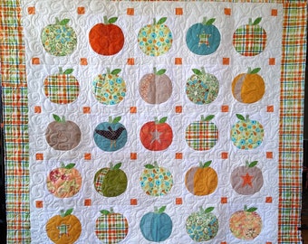 May include: A handmade quilt featuring a repeating pattern of apple shapes in various colors and patterns. The apples are appliqued onto a white background with quilted details. The quilt is bordered with a plaid fabric in orange, green, and white.