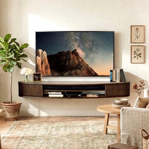 May include: A dark wood floating media console with a curved, slatted design mounted on a wall beneath a flat-screen television displaying a mountain landscape. The console features open shelving holding books and electronic devices in a living room.