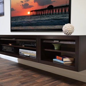 May include: A dark brown, wall-mounted entertainment center with open shelving and a closed cabinet. The unit is mounted below a large flat-screen TV. A decorative white sphere sits on top. The room has wood flooring and a painting of a surfer.