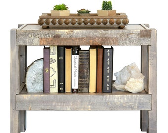 May include: A rustic wooden end table with two shelves. The top shelf has a wooden tray with three small potted succulents. The bottom shelf has a collection of books and a white crystal. The table is painted in a gray wash.