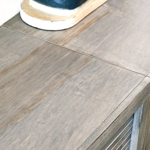 May include: A close-up of a wooden cabinet with a light gray finish. The cabinet has a slatted door and a top surface with visible wood grain. A portion of a white and black shoe is visible on the top.