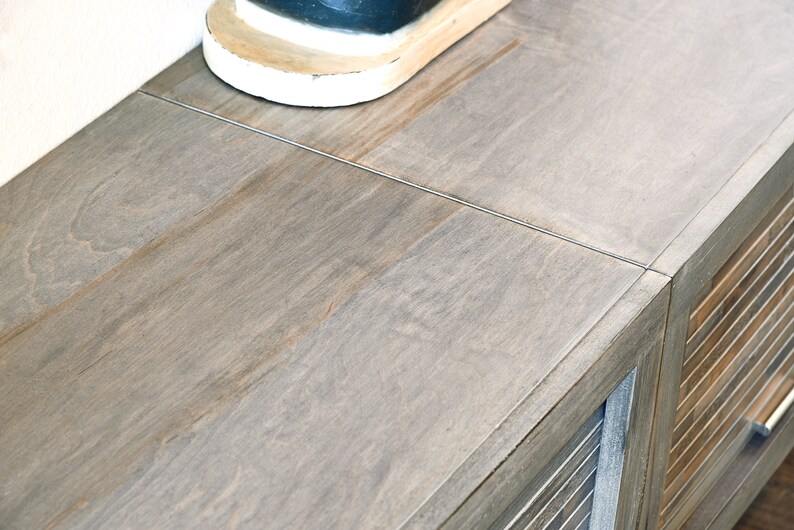 May include: A close-up of a wooden cabinet with a light gray finish. The cabinet has a slatted door and a top surface with visible wood grain. A portion of a white and black shoe is visible on the top.