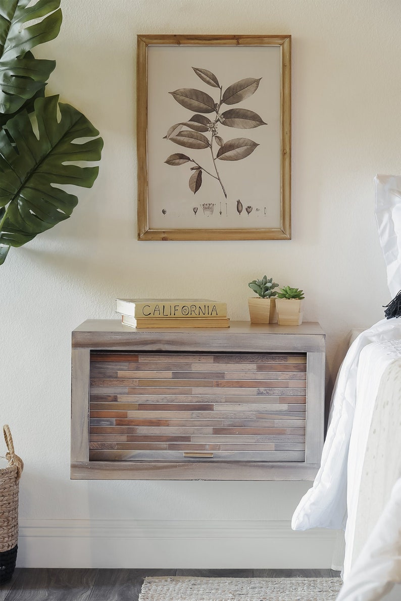 K&ouml;nnte beinhalten: Ein schwebender Nachttisch aus hellem Holz mit einer Schublade und einer Front aus Holzimitat-Laminat. Auf dem Nachttisch stehen zwei B&uuml;cher und zwei kleine Topfpflanzen. Der Nachttisch ist an einer wei&szlig;en Wand neben einem wei&szlig;en Bett mit einer wei&szlig;en Bettdecke befestigt.