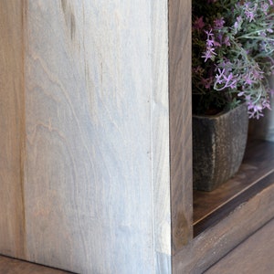 May include: Close-up of a wooden shelf unit with a light gray finish. The corner of the shelf is visible, showing the joinery. A small potted plant with purple flowers sits on the shelf, adding a touch of color.