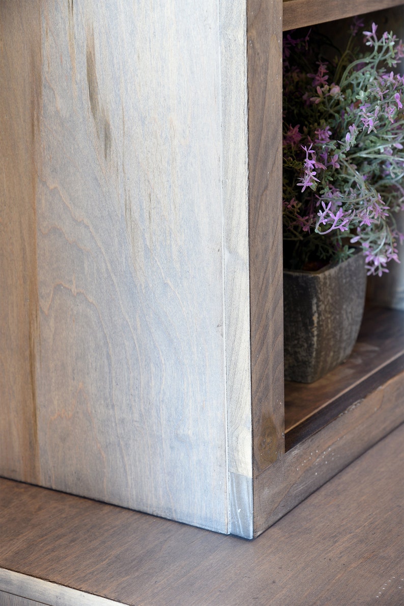 May include: Close-up of a wooden shelf unit with a light gray finish. The corner of the shelf is visible, showing the joinery. A small potted plant with purple flowers sits on the shelf, adding a touch of color.