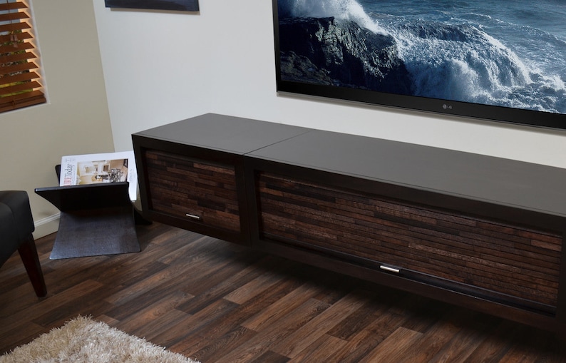 May include: A contemporary, dark brown TV unit with two sliding doors, each with a textured wooden panel design. The unit is wall-mounted, with a large flat-screen TV above. A magazine rack is visible to the left.