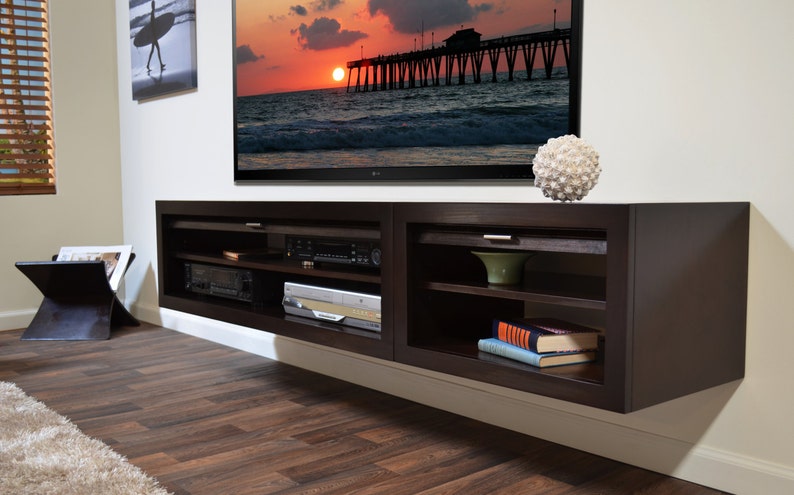 May include: A dark brown, wall-mounted entertainment center with open shelving and a closed cabinet. The unit is mounted below a large flat-screen TV. A decorative white sphere sits on top. The room has wood flooring and a painting of a surfer.