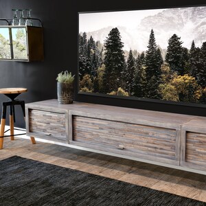 May include: A modern, gray wooden entertainment center with three drawers, featuring a mosaic-style front panel. The unit has a silver base and is positioned below a large flat-screen TV displaying a forest scene. A black rug and a small side table are in the foreground.