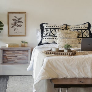 K&ouml;nnte beinhalten: Ein wei&szlig;es Bett mit schwarz-wei&szlig;en Kissen und einem Laptop auf einem Holztablett. Das Bett hat eine wei&szlig;e Bettdecke und eine braune Wolldecke. Neben dem Bett steht ein kleiner Nachttisch aus Holz mit einer Schublade und einer Topfpflanze.