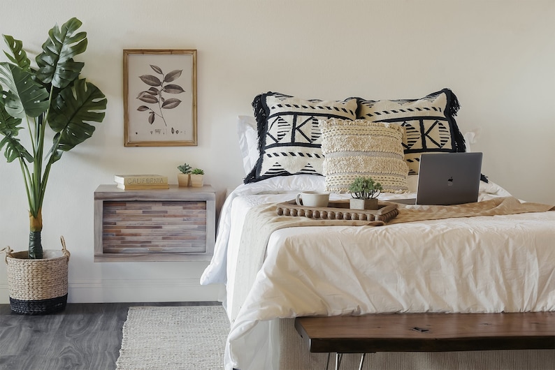 K&ouml;nnte beinhalten: Ein wei&szlig;es Bett mit schwarz-wei&szlig;en Kissen und einem Laptop auf einem Holztablett. Das Bett hat eine wei&szlig;e Bettdecke und eine braune Wolldecke. Neben dem Bett steht ein kleiner Nachttisch aus Holz mit einer Schublade und einer Topfpflanze.