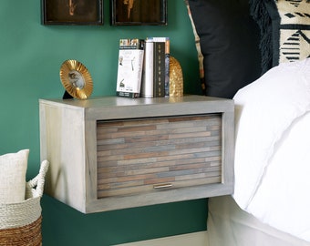 May include: A gray floating nightstand with a wood-look drawer front. The nightstand is mounted on a green wall and has a gold-colored decorative object and books on top.