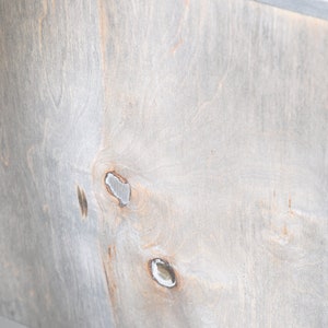 May include: Close-up of a wooden surface with a light gray stain. The wood grain is visible, and there are several knots. A thin metal strip runs along the top edge. The wood has a natural, rustic appearance.
