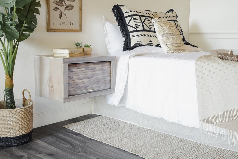 K&ouml;nnte beinhalten: Ein wei&szlig;es Bett mit einer wei&szlig;en Decke und einer braunen Wolldecke. Das Bett hat ein h&ouml;lzernes Kopfteil und einen schwebenden Nachttisch mit einer Schublade. Der Nachttisch ist aus Holz mit einer hellbraunen Oberfl&auml;che. Auf dem Nachttisch stehen zwei kleine Topfpflanzen.