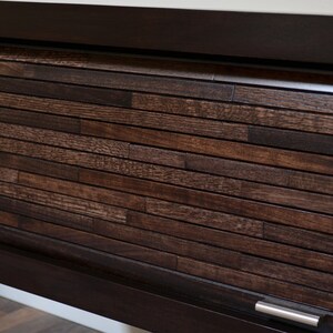 May include: A dark brown wooden cabinet door with a horizontal plank design. The door is framed in a darker wood and features a silver handle. The wood grain is visible, adding texture.