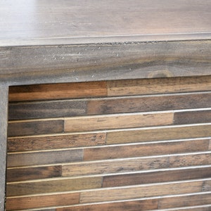 May include: Close-up of a wooden cabinet corner. The top and frame are a muted gray-brown, while the front features a mosaic of horizontal wooden planks in varying shades of brown and tan. The cabinet has a modern, rustic aesthetic.