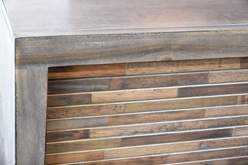 May include: Close-up of a wooden cabinet corner. The top and frame are a muted gray-brown, while the front features a mosaic of horizontal wooden planks in varying shades of brown and tan. The cabinet has a modern, rustic aesthetic.