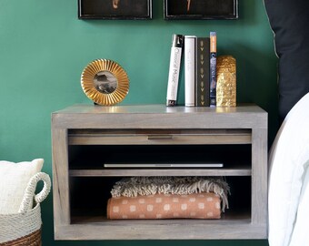 May include: A floating nightstand with two shelves, a drawer, and a gold-toned picture frame. The nightstand is made of wood and has a light brown finish. The shelves are empty, but the drawer is closed. The picture frame is on the top shelf and contains a black and white photo. There are several books and a gold-toned decorative object on the top shelf. The nightstand is mounted on a green wall.