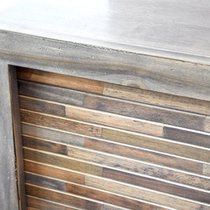 May include: Close-up of a wooden cabinet corner. The cabinet has a gray-stained frame and a front panel made of rectangular wood pieces in shades of brown and tan. The top surface is also gray.