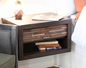 May include: Dark brown floating nightstand with a drawer featuring a mosaic wood design. The nightstand has an open shelf with books. A lamp and a magazine are on top. The nightstand is next to a bed with white bedding.