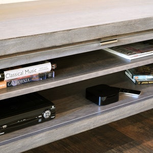 May include: A gray wooden entertainment center with multiple shelves. The shelves hold books, magazines, a DVD player, a streaming device, and a remote control. The top surface is light gray.