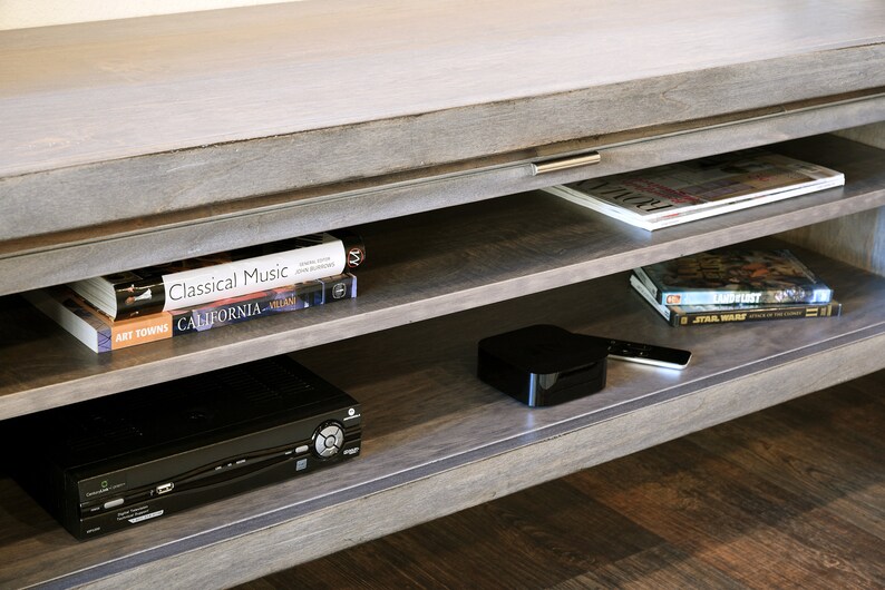 May include: A gray wooden entertainment center with multiple shelves. The shelves hold books, magazines, a DVD player, a streaming device, and a remote control. The top surface is light gray.