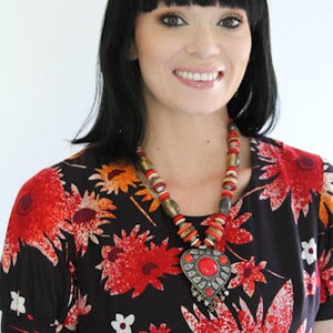 May include: A woman with black hair and bangs is wearing a black and red floral dress with a large silver pendant necklace with red beads. The pendant is shaped like a heart with a red stone in the center.