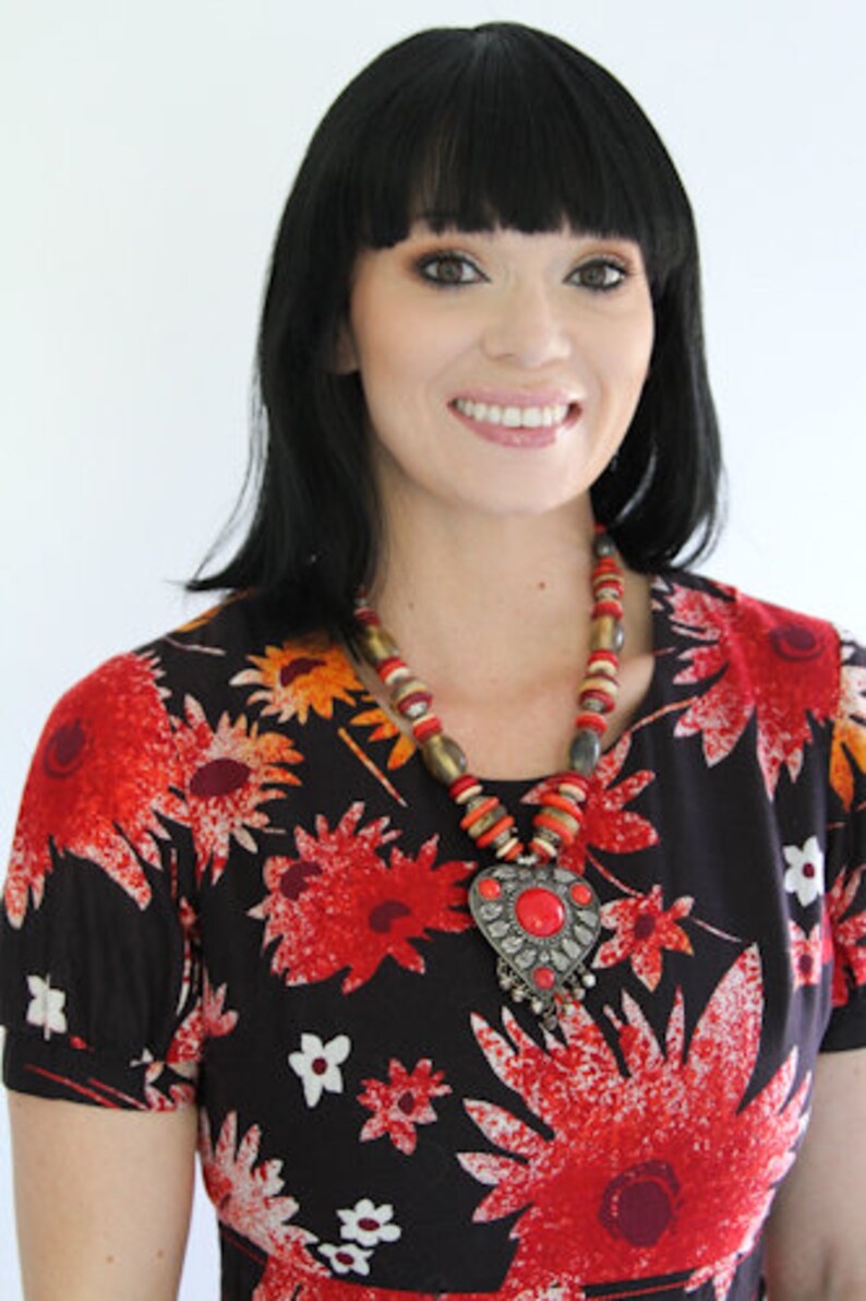 May include: A woman with black hair and bangs is wearing a black and red floral dress with a large silver pendant necklace with red beads. The pendant is shaped like a heart with a red stone in the center.