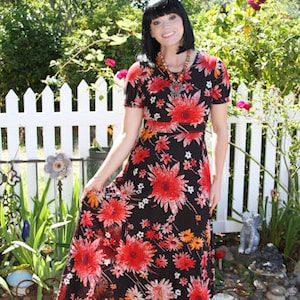 May include: A woman wearing a black maxi dress with a red and orange floral print. The dress has short sleeves and a fitted bodice. She is standing in front of a white picket fence.