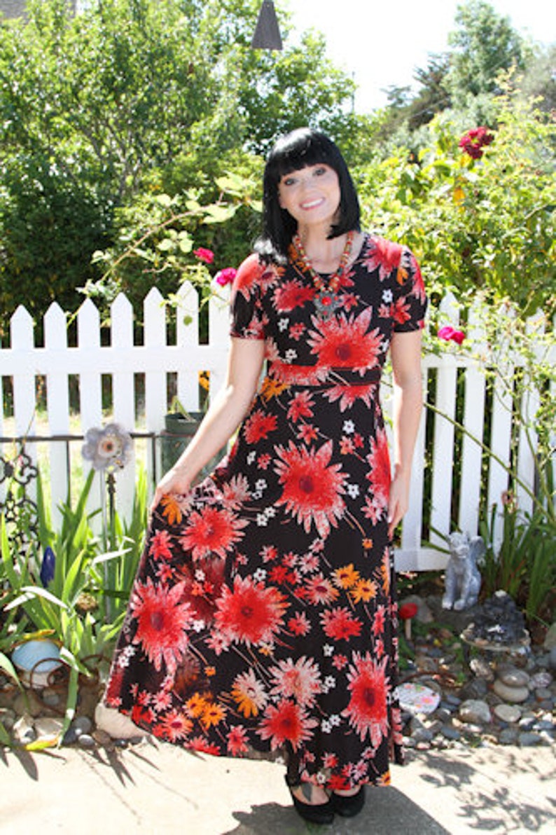 May include: A woman wearing a black maxi dress with a red and orange floral print. The dress has short sleeves and a fitted bodice. She is standing in front of a white picket fence.