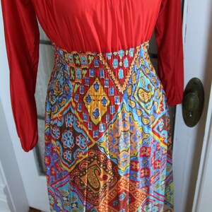 K&ouml;nnte beinhalten: Ein langes rotes und mehrfarbiges Maxikleid mit geometrischem Muster. Das Kleid hat einen figurbetonten Oberteil und einen flie&szlig;enden Rock.