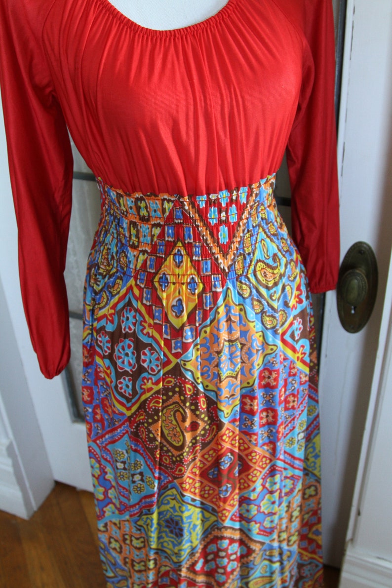 K&ouml;nnte beinhalten: Ein langes rotes und mehrfarbiges Maxikleid mit geometrischem Muster. Das Kleid hat einen figurbetonten Oberteil und einen flie&szlig;enden Rock.
