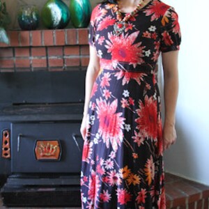 May include: A woman wearing a long, black maxi dress with a floral print. The dress has red, orange, and white flowers on a black background. She is wearing a long necklace with a large pendant.