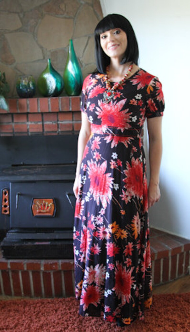 May include: A woman wearing a long, black maxi dress with a floral print. The dress has red, orange, and white flowers on a black background. She is wearing a long necklace with a large pendant.