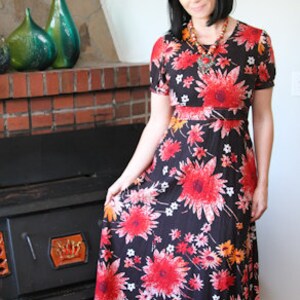 May include: A woman wearing a long, black maxi dress with a red, orange, and white floral print. The dress has a fitted bodice and a flowing skirt. She is standing in front of a fireplace.