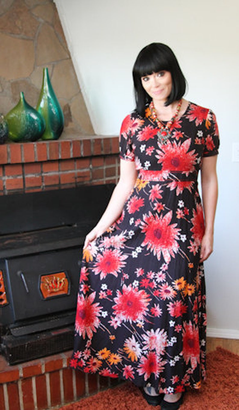 May include: A woman wearing a long, black maxi dress with a red, orange, and white floral print. The dress has a fitted bodice and a flowing skirt. She is standing in front of a fireplace.