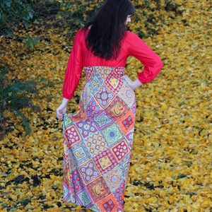 K&ouml;nnte beinhalten: Eine Frau tr&auml;gt ein rotes Maxikleid mit langen &Auml;rmeln und einem mehrfarbigen Muster. Sie steht auf einem Feld mit gelben Bl&auml;ttern.