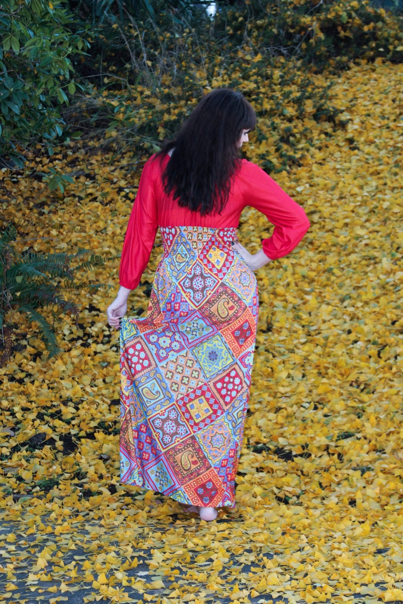 K&ouml;nnte beinhalten: Eine Frau tr&auml;gt ein rotes Maxikleid mit langen &Auml;rmeln und einem mehrfarbigen Muster. Sie steht auf einem Feld mit gelben Bl&auml;ttern.