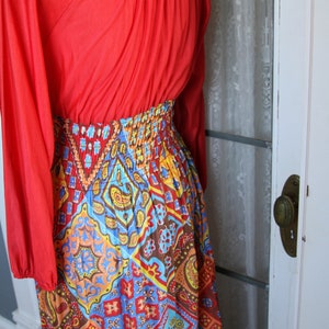 K&ouml;nnte beinhalten: Ein langes rotes Kleid mit mehrfarbigem Muster und einer gerafften Taille. Das Kleid hat lange &Auml;rmel und einen runden Ausschnitt.