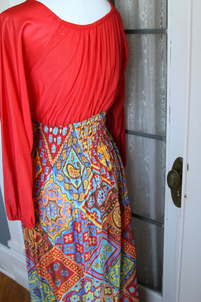 K&ouml;nnte beinhalten: Ein langes rotes Kleid mit mehrfarbigem Muster und einer gerafften Taille. Das Kleid hat lange &Auml;rmel und einen runden Ausschnitt.