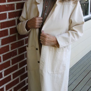 May include: A white long-sleeved jacket with a button closure and two pockets. The jacket has a blue and green fish design on the bottom. The model is wearing blue jeans.