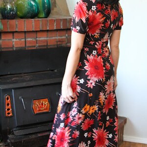 May include: A woman wearing a long, black maxi dress with a floral print. The dress has red, orange, and white flowers on a black background. The woman is standing in front of a fireplace.