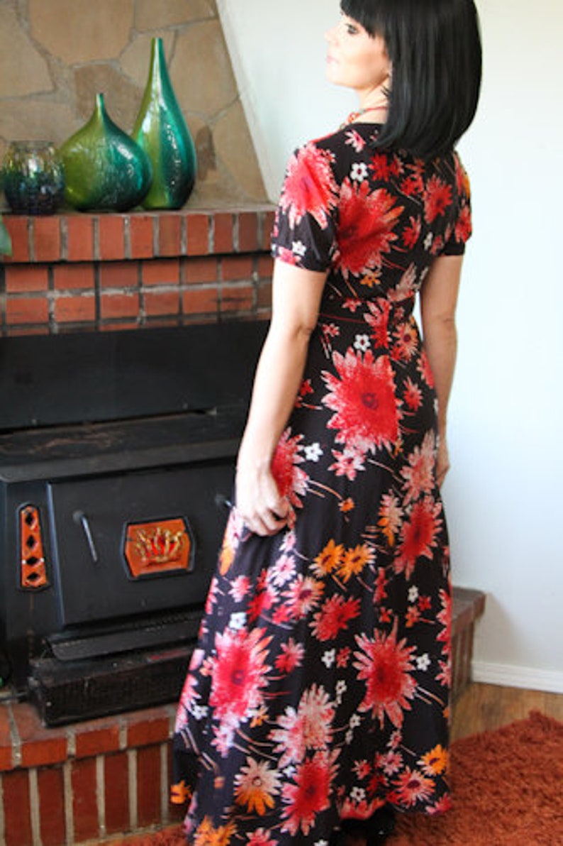May include: A woman wearing a long, black maxi dress with a floral print. The dress has red, orange, and white flowers on a black background. The woman is standing in front of a fireplace.
