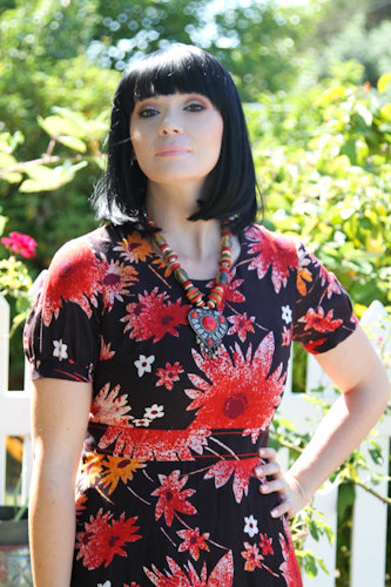 May include: A woman with black hair is wearing a black and red floral dress with a long, beaded necklace with a large, ornate pendant.