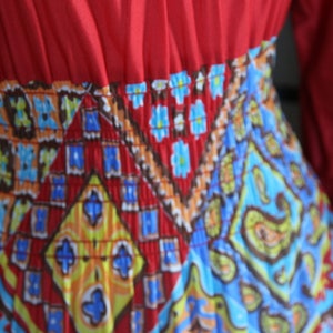 K&ouml;nnte beinhalten: Ein rotes Kleid mit einem bunten geometrischen Muster. Das Kleid hat ein tailliertes Oberteil und einen flie&szlig;enden Rock.